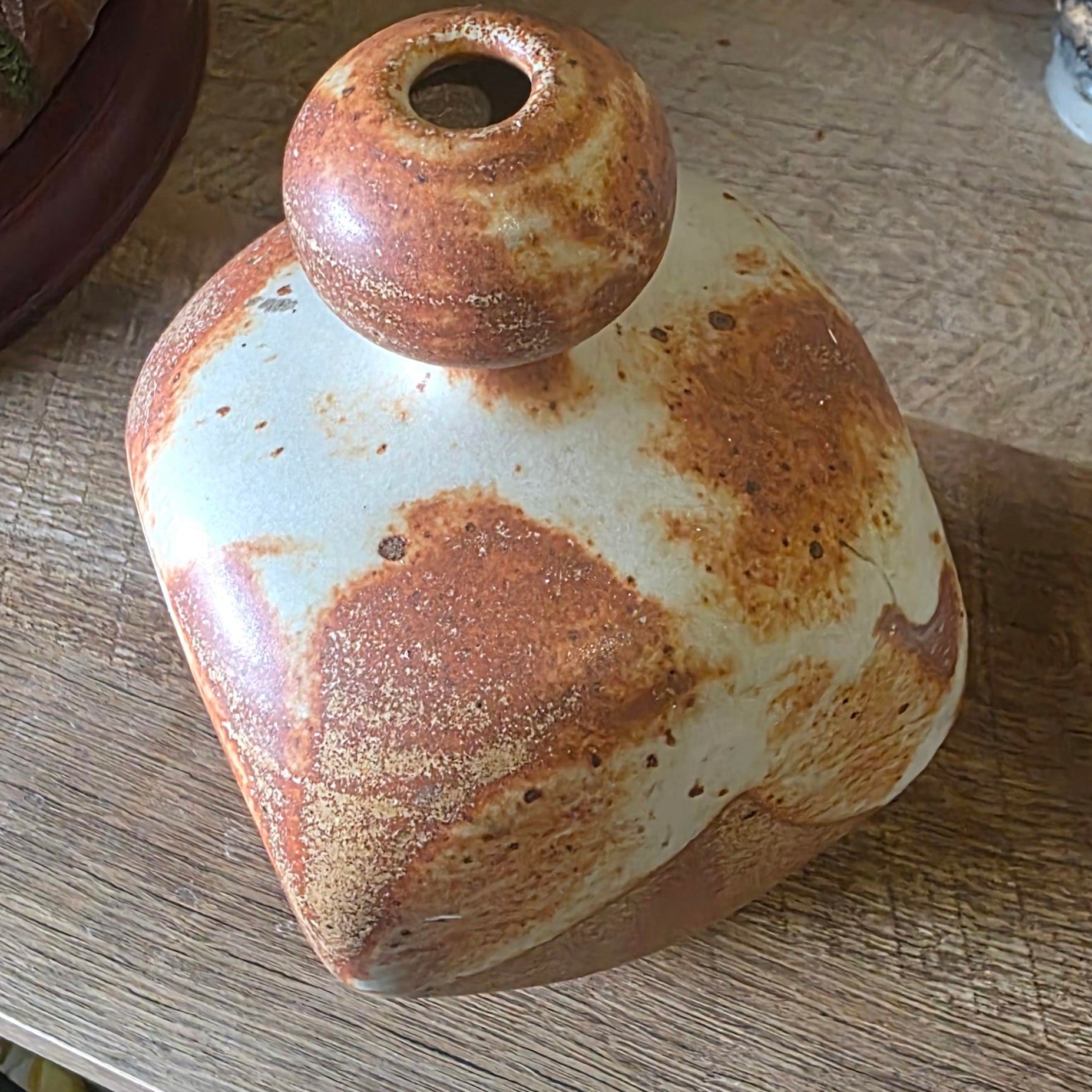 A rustic handmade ceramic bottle or vase with a rounded body, a small neck opening, and a wide flared base, featuring a white glaze with brown rust-like mottling, displayed on a wooden tabletop.