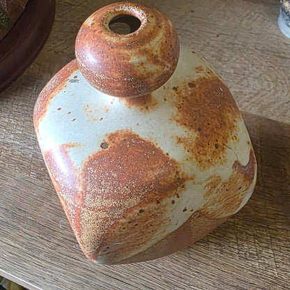 A rustic handmade ceramic bottle or vase with a rounded body, a small neck opening, and a wide flared base, featuring a white glaze with brown rust-like mottling, displayed on a wooden tabletop.