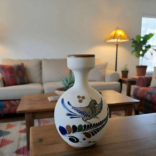 A white ceramic vase with a hand-painted bird and leaf pattern, displayed on a wooden table in a cozy living room setting.